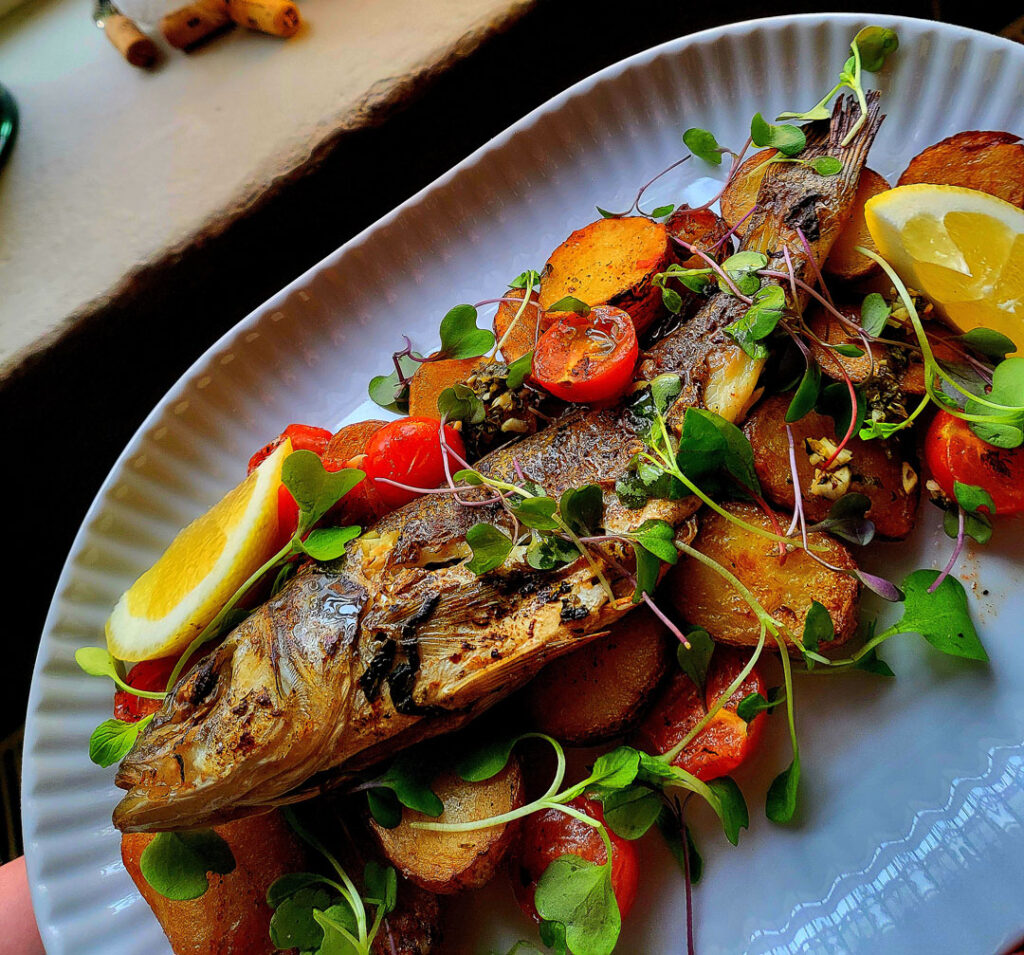 Kokonainen paistettu kala Bistro Wool Factoryssa Hyvinkäällä, tarjoiltuna paahdettujen perunoiden, kirsikkatomaattien, sitruunan ja tuoreiden versojen kanssa valkoisella lautasella. Whole roasted fish at Bistro Wool Factory in Hyvinkää, served with roasted potatoes, cherry tomatoes, lemon wedges and fresh microgreens on a white plate.