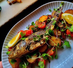 Kokonainen paistettu kala Bistro Wool Factoryssa Hyvinkäällä, tarjoiltuna paahdettujen perunoiden, kirsikkatomaattien, sitruunan ja tuoreiden versojen kanssa valkoisella lautasella. Whole roasted fish at Bistro Wool Factory in Hyvinkää, served with roasted potatoes, cherry tomatoes, lemon wedges and fresh microgreens on a white plate.